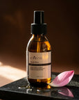 Evera Liquid Gloss Treatment No 0 with lotus petal and oil droplets on charcoal stone, deep umber editorial backdrop